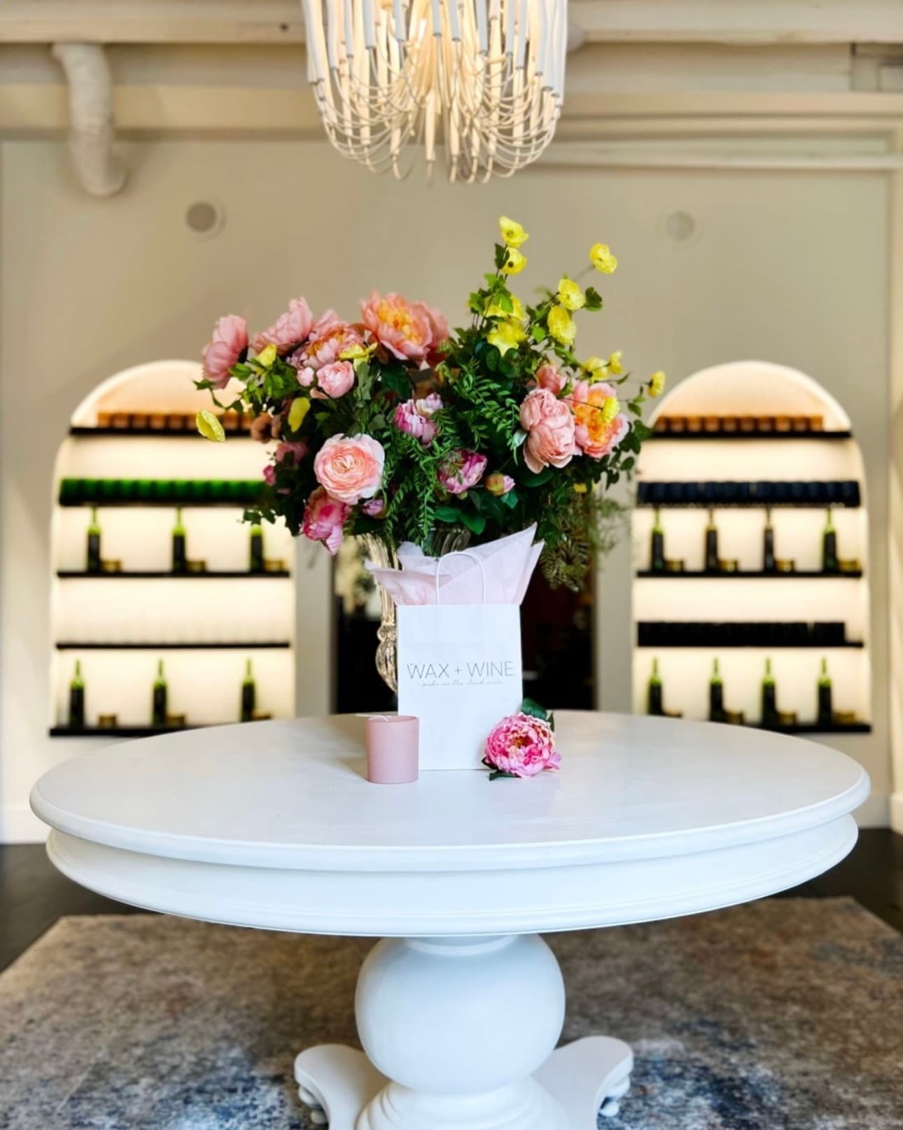 A pedestal table with lush pink and coral florals and a Wax + Wine bag, set in front of the Scent Cellar