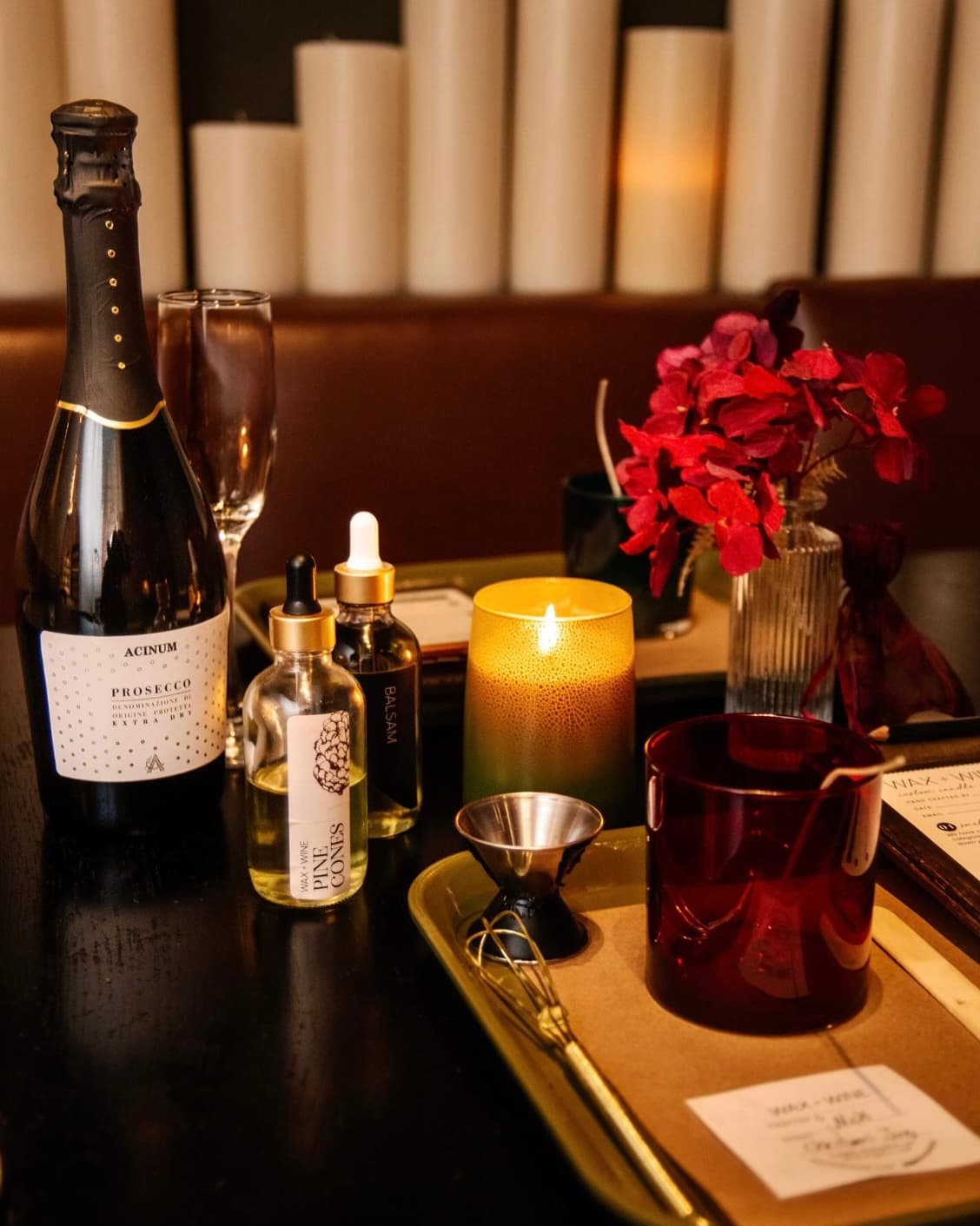 A bottle of prosecco with a Wax + Wine candle kit and red florals on a dark table